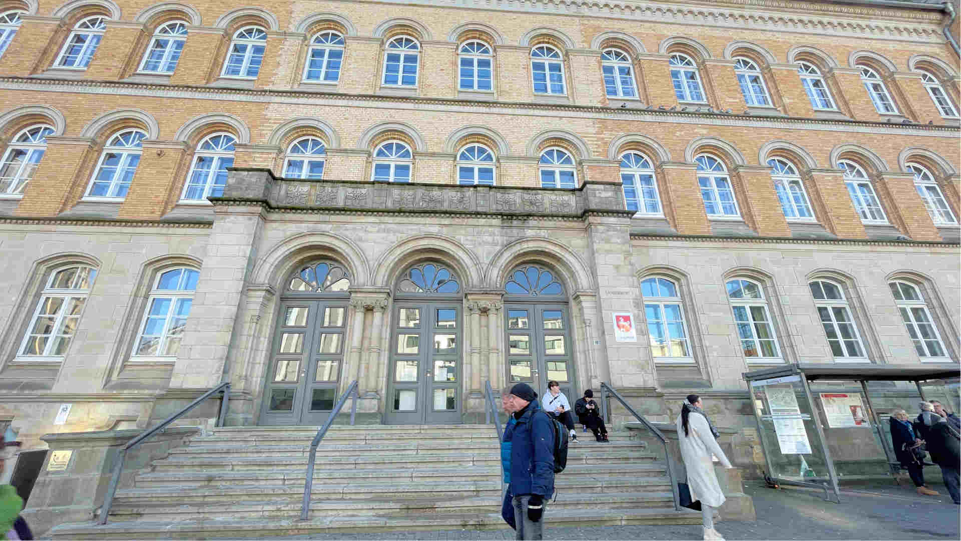 Der Eingang vom Landgericht Osnabrück. Präsent mittig ist das Eingangsportal mit den Stufen, drum herum ist noch etwas vom Gebäude abgebildet. Der Ausschnitt reicht bis zum zweiten Stock. Rechts im Bild ist das Bushaltestellenhäuschen des Neumarkts. Vor dem Gebäude laufen Menschen.